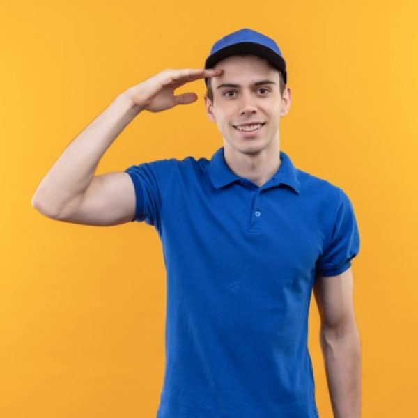 young courier wearing blue uniform and blue cap doing happy honor over yellow isolated background